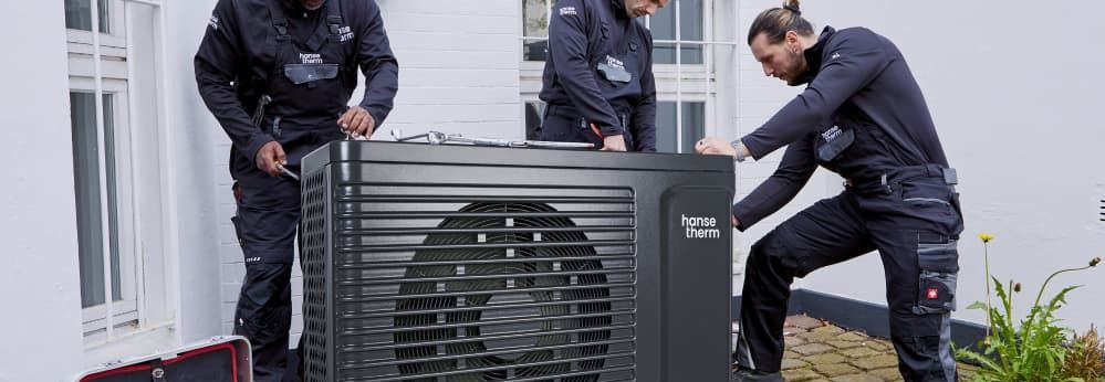 Drei Personen installieren eine Wärmepumpe. Das Logo der Marke hansetherm ist abgebildet.